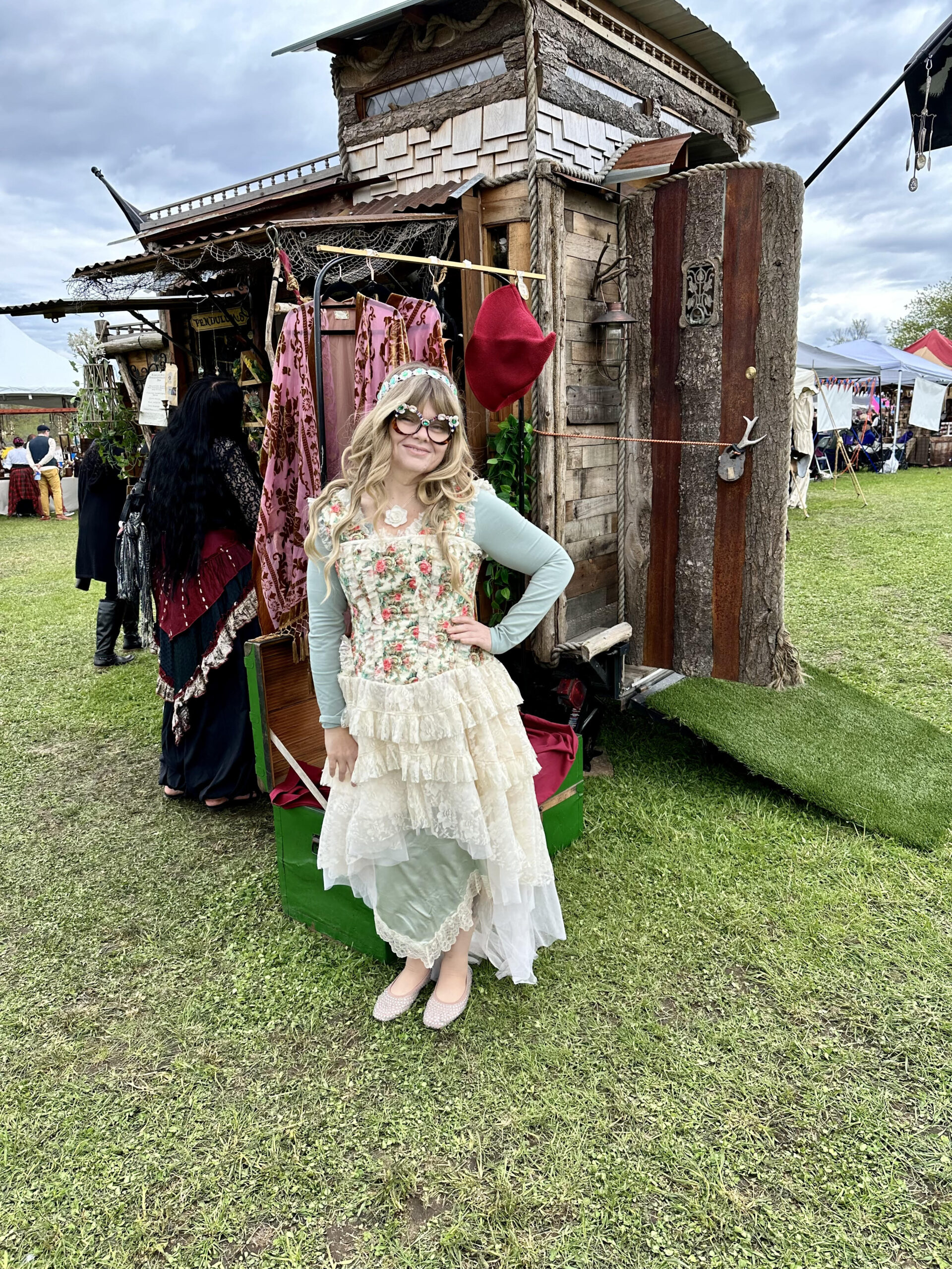 Photo of Nikki, 'The Glittered Witch,' at a Renaissance Faire, dressed in light, airy attire. She wears a pastel floral embroidered blouse and a layered, multi-tiered skirt in shades of light green and white lace. Her large, round glasses have ornate frames, complementing her soft, styled look. Nikki stands confidently in front of a rustic wooden booth adorned with colorful textiles. The scene captures a festive and historical ambiance.