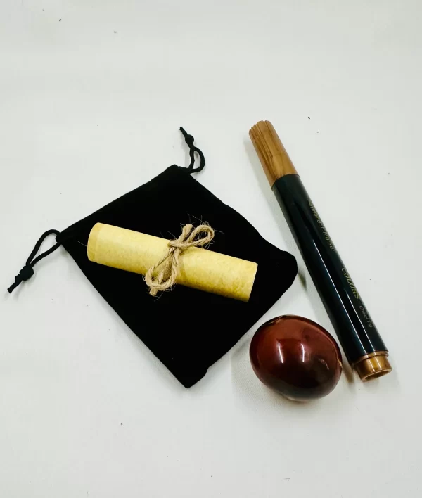 Photo of a 'Make Your Own Sigil Kit' including a polished, oval-shaped carnelian tubule, a gold-tone marker with a wooden-like finish, and rolled yellow parchment tied with twine. These items are displayed alongside a black velvet pouch with a drawstring closure, all laid out on a plain white background to highlight each component of the kit.