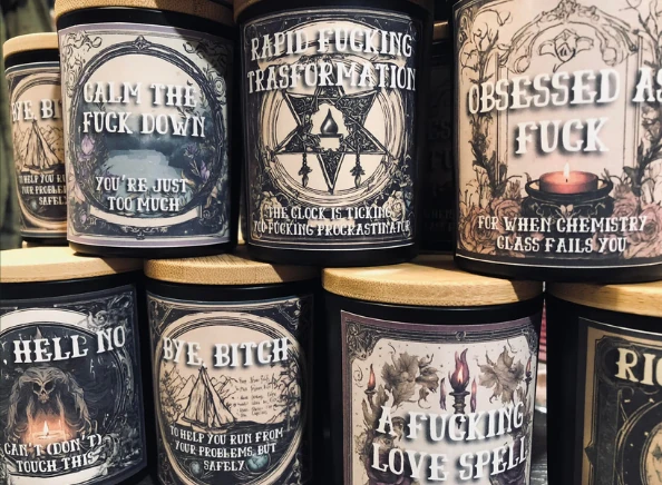 A display of candles with labels such as "Calm the Fuck Down", "Rapid Fucking Transformation", "Obsessed As Fuck", "Oh Hell No", "Bye Bitch", and "A Fucking Love Spell"