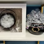 A Halloween crafting kit containing a black cauldron, a white box with faux smoke and bubbles. Next to it is an example completed cauldrom