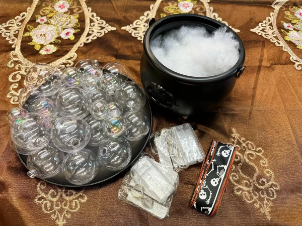 A Halloween cauldron centerpiece craft kit. The kit includes a black cauldron filled with white faux cotton, a tray of clear bubble ornaments, battery-powered lights, and a skeleton ribbon.