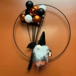 A Halloween wreath made with a wire hoop decorated with black, orange, and white balloons. A small gnome wearing a witch's hat is attached to the wreath.