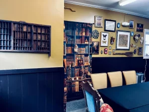 The image shows our cozy crafting space with large wooden tables in the center, inviting people to gather and create. The room is filled with warm lighting and a rustic, homey ambiance. In the background, what may appear to be a bookshelf is actually a curtain with decorative elements on it, adding texture and a whimsical touch to the space. The area is surrounded by shelves filled with crafting supplies and mystical items, creating an inspiring and welcoming environment for creative activities and group projects.