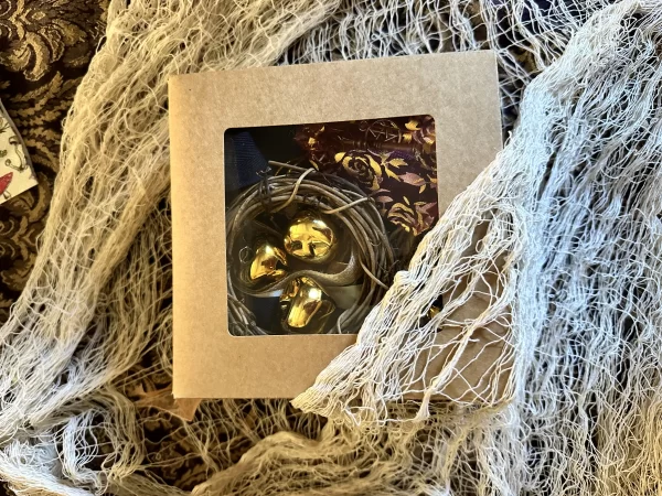 Witch bell kit packaged in a craft box with a clear window, showing gold bells and other crafting materials, surrounded by lace fabric for a rustic presentation.