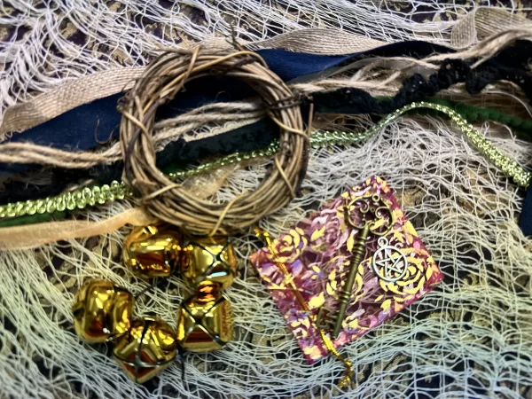 Materials for making witch bells, including gold bells, a vine wreath, various ribbons, a decorative key, and charms, arranged on textured lace fabric.