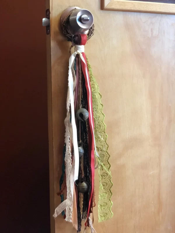 Handmade witch bells hanging on a doorknob, featuring a vine wreath and various colored ribbons with bells attached, used for spiritual cleansing and decoration.