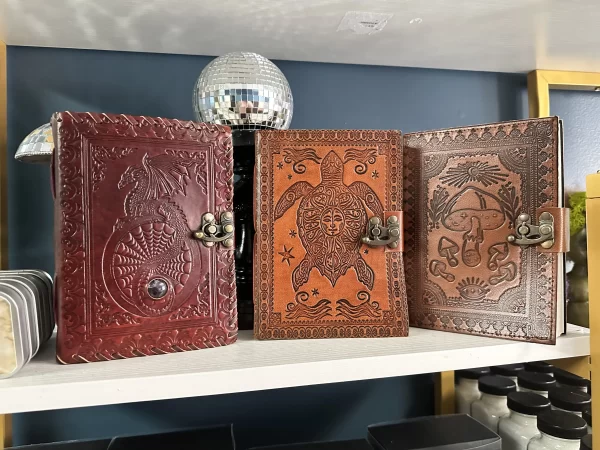 Three embossed leather journals with dragon, turtle, and mushroom designs on a shelf at Broomstick Adventures, featuring antique-style clasps.