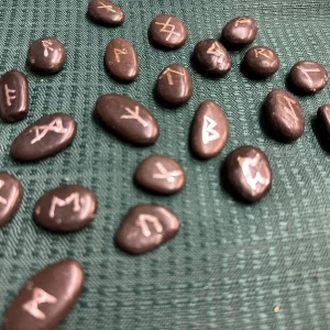 Handmade rune stones displayed on a textured green fabric, featuring various symbols that participants will learn to create in the 'Make Your Own Runes' class.