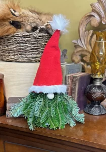 Christmas gnome decoration made of fresh pine branches, featuring a red hat with a fluffy white trim and pom-pom, displayed on a wooden surface with vintage books and decor in the background.