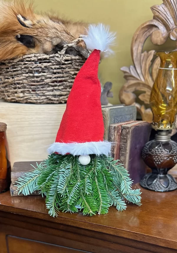 Christmas gnome decoration made of fresh pine branches, featuring a red hat with a fluffy white trim and pom-pom, displayed on a wooden surface with vintage books and decor in the background.