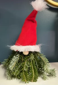 Christmas gnome decoration made of fresh pine branches, featuring a red hat with white trim and a fluffy pom-pom, set against a dark teal background.