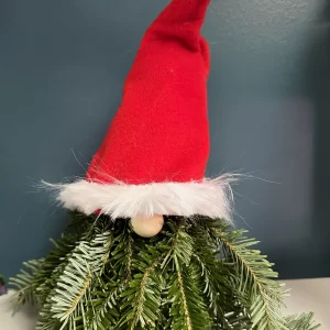 Christmas gnome decoration made of fresh pine branches, featuring a red hat with white trim and a fluffy pom-pom, set against a dark teal background.