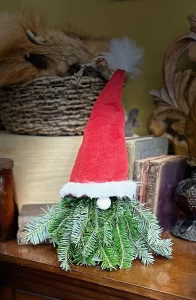 Christmas gnome decoration crafted from fresh pine branches, topped with a red Santa hat featuring white trim and a fluffy pom-pom, displayed on a wooden surface with vintage books and decor in the background.