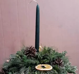 A handcrafted Yule centerpiece featuring lush greenery, pinecones, dried orange slices, and a tall green taper candle at the center. The arrangement is set against a simple pinkish background, showcasing its festive and natural holiday aesthetic.