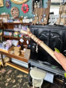 A handcrafted wand held in focus, featuring a wooden shaft wrapped in thin red wire with a small key charm hanging near the handle. The tip of the wand is adorned with a purple crystal. The background is a cozy, eclectic shop filled with shelves of mystical and decorative items, including colorful crystals, jewelry, and other magical trinkets. The scene conveys a whimsical and magical atmosphere.