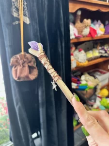 A handcrafted wand held in focus, featuring a wooden shaft wrapped in thin red wire with a small key charm hanging near the handle. The tip of the wand is adorned with a purple crystal. The background is a cozy, eclectic shop filled with shelves of mystical and decorative items, including colorful crystals, jewelry, and other magical trinkets. The scene conveys a whimsical and magical atmosphere.