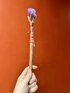 A handcrafted wand held upright against an orange background. The wand features a natural wooden shaft wrapped with copper wire, a small dangling key charm, and a purple crystal securely affixed at the tip. The minimalist background highlights the intricate details of the wand, emphasizing its rustic and magical design.