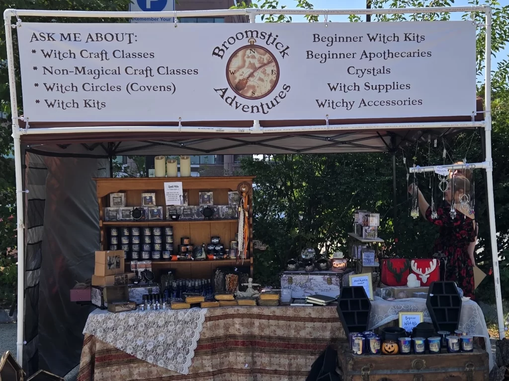 Outdoor market booth for Broomstick Adventures featuring a wide banner listing witchcraft classes and supplies, a wooden shelf of candles and kits, and a decorated table with crystals, journals, and accessories.